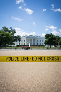 A prominent government building stands in the background, partially obscured by a bright yellow police line with the words 'POLICE LINE - DO NOT CROSS'. The sky is clear with some scattered clouds, and there are trees flanking the building.