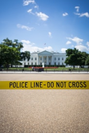 A prominent government building stands in the background, partially obscured by a bright yellow police line with the words 'POLICE LINE - DO NOT CROSS'. The sky is clear with some scattered clouds, and there are trees flanking the building.