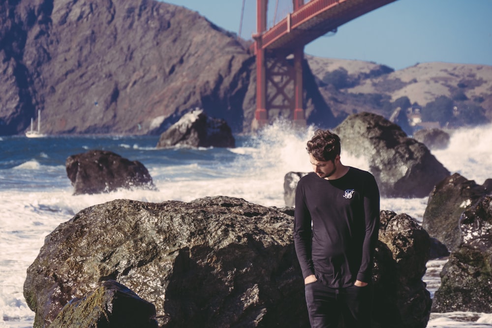 man standing beside black stone with sea waves near red metal bridge during daytime