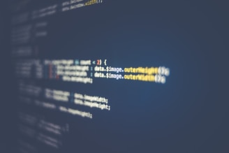 A close-up view of a computer screen displaying lines of code. The code features syntax elements like variables, functions, and brackets, highlighted in different colors against a dark background. The text is slightly out of focus along the edges, with a more focused section in the middle.