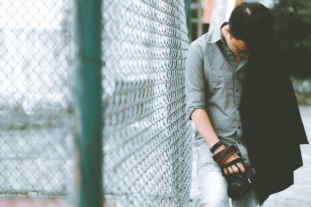 man leaning on cyclone fence while holding DSLR camera