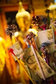The image depicts a statue of Buddha in the background with a focus on various banknotes attached to sticks, adorned with colorful ribbons. The setting appears to be a temple or a place of worship, with a warm, golden ambiance.