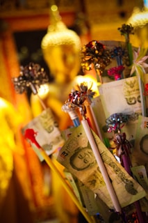 The image depicts a statue of Buddha in the background with a focus on various banknotes attached to sticks, adorned with colorful ribbons. The setting appears to be a temple or a place of worship, with a warm, golden ambiance.