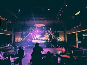 A dimly lit nightclub interior with neon lighting and geometric designs on the stage. Heart-shaped light patterns are visible, along with a large neon sign that reads 'Kitty Palace.' The room is filled with tables and chairs, and a few silhouettes of people can be seen in the foreground.