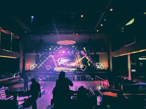 A dimly lit nightclub interior with neon lighting and geometric designs on the stage. Heart-shaped light patterns are visible, along with a large neon sign that reads 'Kitty Palace.' The room is filled with tables and chairs, and a few silhouettes of people can be seen in the foreground.