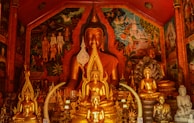 A richly decorated room filled with multiple golden Buddha statues of various sizes. The central large Buddha statue is framed by elaborate murals featuring religious and historical scenes. The warm lighting accentuates the gold tones and intricate details carved into the statues.