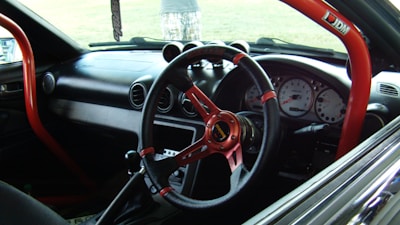 A car interior featuring a racing-style steering wheel with red accents and a roll cage bar. The dashboard includes several gauges, and an air freshener is hanging from the rearview mirror. The image captures the front passenger area with a clear view through the windshield.