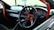 A car interior featuring a racing-style steering wheel with red accents and a roll cage bar. The dashboard includes several gauges, and an air freshener is hanging from the rearview mirror. The image captures the front passenger area with a clear view through the windshield.