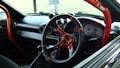 A car interior featuring a racing-style steering wheel with red accents and a roll cage bar. The dashboard includes several gauges, and an air freshener is hanging from the rearview mirror. The image captures the front passenger area with a clear view through the windshield.