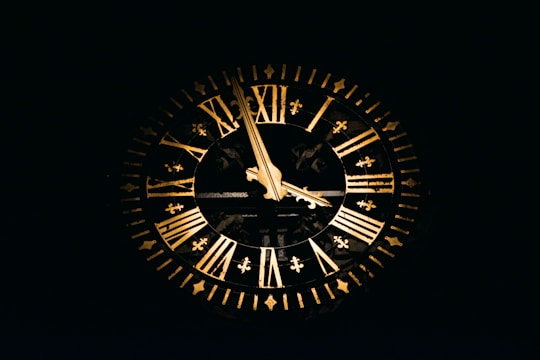 A large, ornate clock with Roman numerals is prominently displayed against a dark background. The clock features two gold-colored hands pointing at different numbers and is adorned with intricate designs along the rim.