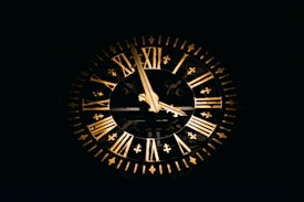 A large, ornate clock with Roman numerals is prominently displayed against a dark background. The clock features two gold-colored hands pointing at different numbers and is adorned with intricate designs along the rim.