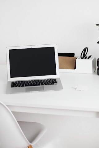 A minimalist workspace featuring a laptop with a black screen on a white desk. On the right side of the desk is an organizer holding scissors, a small cardboard box, and other stationery items. A white chair is partially visible in the foreground, and a potted plant can be seen on the far right of the desk.
