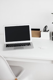 A minimalist workspace featuring a laptop with a black screen on a white desk. On the right side of the desk is an organizer holding scissors, a small cardboard box, and other stationery items. A white chair is partially visible in the foreground, and a potted plant can be seen on the far right of the desk.