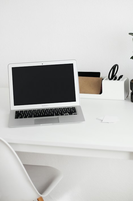 A minimalist workspace featuring a laptop with a black screen on a white desk. On the right side of the desk is an organizer holding scissors, a small cardboard box, and other stationery items. A white chair is partially visible in the foreground, and a potted plant can be seen on the far right of the desk.