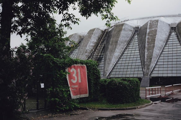 A large, dome-shaped structure with triangular glass panel sections. Overgrown greenery and bushes partially cover the foreground. A red banner with the number 31 and the word 'Tennis' in both Russian and English is visible, hanging on a fence.