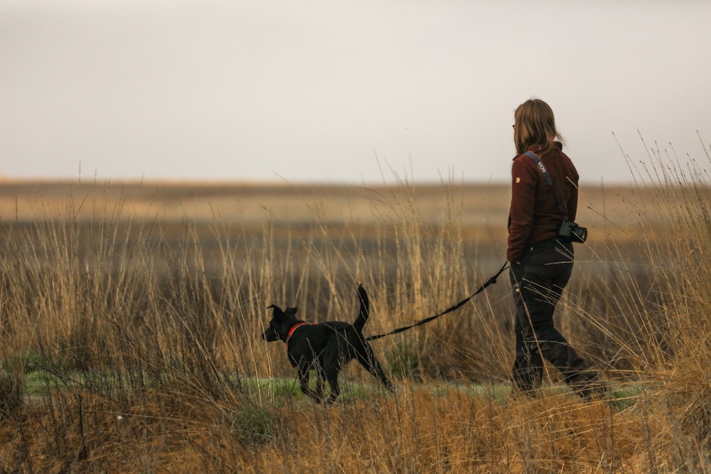 woman in brown jacket and black pants walking with black labrador retriever on brown grass field