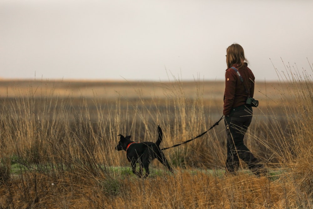 Frau in brauner Jacke und schwarzer Hose geht mit schwarzem Labrador Retriever auf braunem Grasfeld spazieren