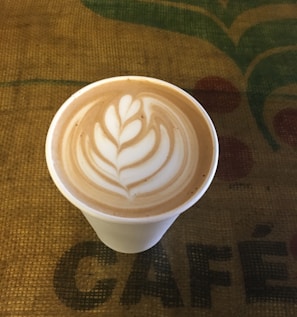 A cup of coffee with intricate latte art featuring a leaf design on the surface, placed on a textured burlap surface with the word 'CAFÉ' partially visible.