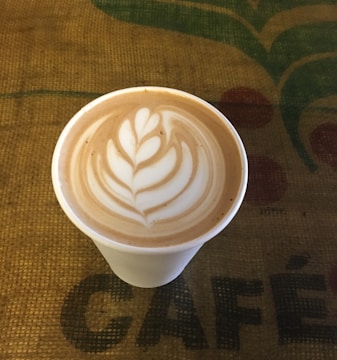 A cup of coffee with intricate latte art featuring a leaf design on the surface, placed on a textured burlap surface with the word 'CAFÉ' partially visible.
