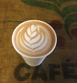 A cup of coffee with intricate latte art featuring a leaf design on the surface, placed on a textured burlap surface with the word 'CAFÉ' partially visible.