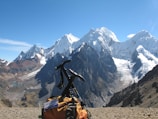 A snow-capped mountain range towers majestically under a clear blue sky, with a notable glacier visible on the peaks. In the foreground, a climbing backpack rests on rocky terrain, with an ice axe attached, suggesting a trekking expedition.