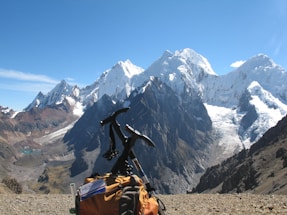 A snow-capped mountain range towers majestically under a clear blue sky, with a notable glacier visible on the peaks. In the foreground, a climbing backpack rests on rocky terrain, with an ice axe attached, suggesting a trekking expedition.