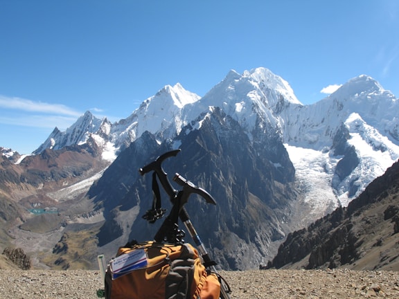 A snow-capped mountain range towers majestically under a clear blue sky, with a notable glacier visible on the peaks. In the foreground, a climbing backpack rests on rocky terrain, with an ice axe attached, suggesting a trekking expedition.