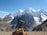 A snow-capped mountain range towers majestically under a clear blue sky, with a notable glacier visible on the peaks. In the foreground, a climbing backpack rests on rocky terrain, with an ice axe attached, suggesting a trekking expedition.