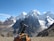 A snow-capped mountain range towers majestically under a clear blue sky, with a notable glacier visible on the peaks. In the foreground, a climbing backpack rests on rocky terrain, with an ice axe attached, suggesting a trekking expedition.