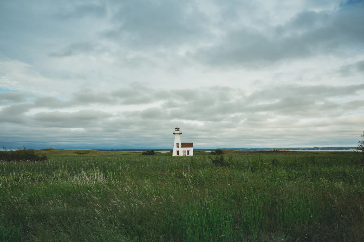 A historic white PEI lighthouse with red trim standing in a green field with tall grass, the ocean horizon visible beyond and an overcast maritime sky above