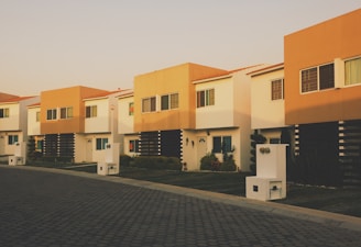 A row of modern two-story houses with flat roofs, characterized by a combination of white and tan exteriors. Each house features a front yard with minimal landscaping, including small bushes and a lawn. The pavement and sidewalks are paved with dark cobblestones, adding a neat and organized feel to the environment. The lighting suggests a time around sunset, casting a warm hue on the buildings.