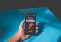 A hand is holding a smartphone displaying the time as 10:22 on a Thursday, August 31. A notification is visible on the screen. The background consists of a smooth surface with a vibrant blue color, partially in shadow.