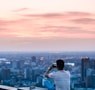 person sitting on edge of building taking photo of city during daytime