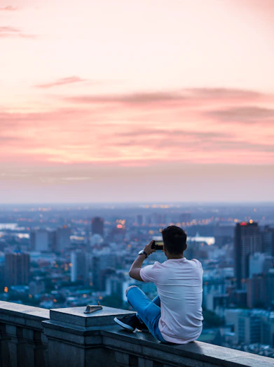 person sitting on edge of building taking photo of city during daytime. Cut the sky litle bit