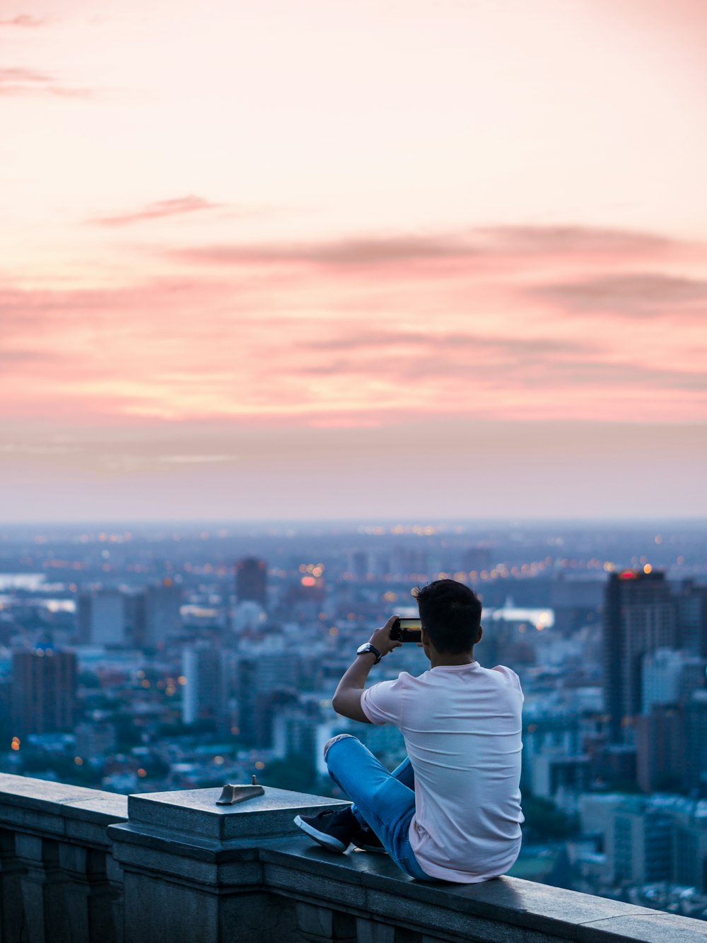 person sitting on edge of building taking photo of city during daytime