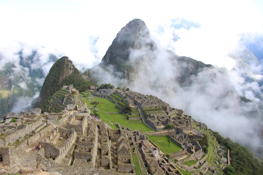 An ancient city with stone structures and terraces set high in the Andes mountains. Clouds partially obscure the dramatic mountain peaks in the background.