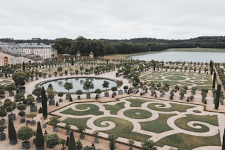 An elaborately designed formal garden features intricate patterns of manicured hedges, geometric shapes, and pathways. The garden is expansive, with a large rectangular pond surrounded by potted citrus trees meticulously arranged. In the background, a grand building with classical architecture is visible, set against a backdrop of lush greenery and trees.