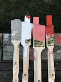 Four paintbrushes with varying paint colors—grey, white, pink, and red—are lined up on a wooden surface. The brushes have paint splatters on their handles, and the bristles are dipped in corresponding colors.