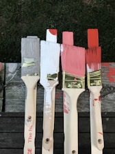 Four paintbrushes with varying paint colors—grey, white, pink, and red—are lined up on a wooden surface. The brushes have paint splatters on their handles, and the bristles are dipped in corresponding colors.