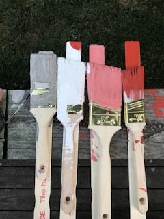 Four paintbrushes with varying paint colors—grey, white, pink, and red—are lined up on a wooden surface. The brushes have paint splatters on their handles, and the bristles are dipped in corresponding colors.