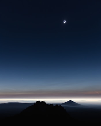 A total solar eclipse dominates the sky, with the sun's corona visible as a halo around the moon. The foreground includes a silhouetted mountain landscape and a clear horizon. Soft hues of blue, orange, and black create a stunning gradient from the top of the sky to the horizon.