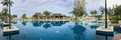 A luxurious, expansive swimming pool surrounded by lush greenery and palm trees, with a scenic sky above. The pool reflects the surrounding trees and a stylish building with a thatched roof, suggesting a resort setting.