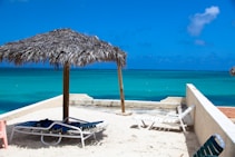A tranquil beach scene with clear turquoise water and a bright blue sky. The foreground features a sandy area with a thatched umbrella providing shade to two lounge chairs. The view is framed by a low wall, and a small hint of pink furniture is visible to the side.