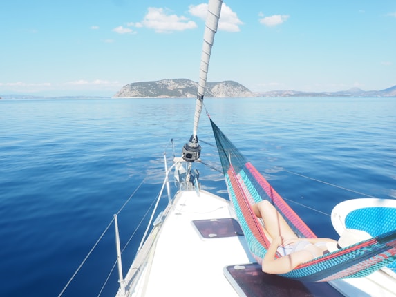 A person relaxing in a colorful hammock on the deck of a boat, surrounded by calm, clear blue water. The horizon features distant hills and a few scattered clouds in a bright, clear sky.