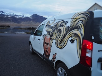 A van parked on a road features a large, detailed graffiti artwork on its side. This artwork includes a stylized portrait of a person with exaggerated, swirling yellow hair. In the background, there are snow-capped mountains and a partly cloudy sky, creating a dramatic natural setting.