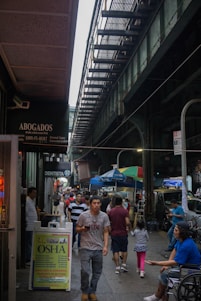 A bustling urban street scene under an elevated train track, with people walking along the sidewalk. Various signs in Spanish advertise services such as legal assistance and dentistry. Street vendors with colorful awnings and people eating add to the lively atmosphere of the city.