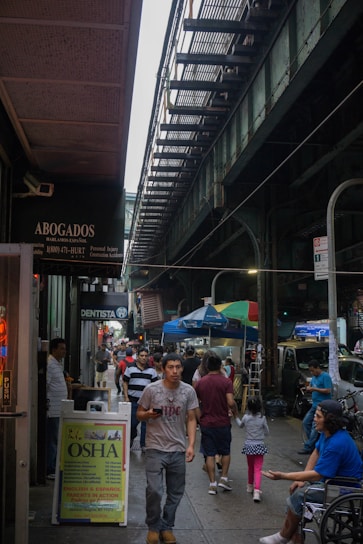 A bustling urban street scene under an elevated train track, with people walking along the sidewalk. Various signs in Spanish advertise services such as legal assistance and dentistry. Street vendors with colorful awnings and people eating add to the lively atmosphere of the city.