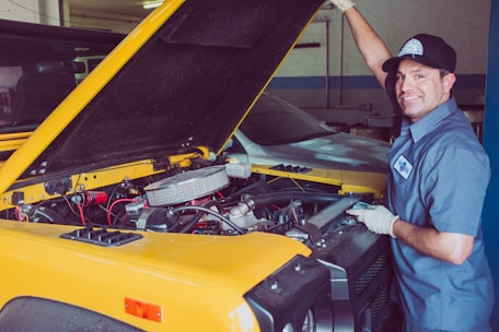 A mechanic is standing next to a yellow car with the hood open. He is smiling and wearing a blue mechanic uniform with a cap and gloves. The car is stationary inside a workshop with another vehicle in the background.