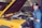 A mechanic is standing next to a yellow car with the hood open. He is smiling and wearing a blue mechanic uniform with a cap and gloves. The car is stationary inside a workshop with another vehicle in the background.