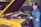 A mechanic is standing next to a yellow car with the hood open. He is smiling and wearing a blue mechanic uniform with a cap and gloves. The car is stationary inside a workshop with another vehicle in the background.